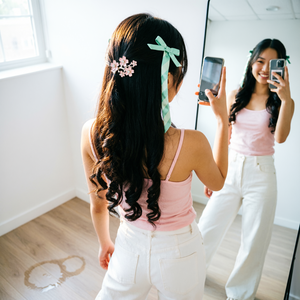 可爱吸睛的发饰，精致的发夹或发带点缀，柔和的自然光线从上方或侧面洒下，营造温暖柔和的光影效果。背景为简约清新，地板纹理清晰，带有水渍，增加细节感，色调清新淡雅，突出青春甜美氛围。采用高位俯拍角度，从上方向下拍摄，强调女孩背影和反射镜中的自拍动作，光线柔和，阴影细腻，增强立体感。人物动作自然，腰背曲线优美，脚踩地面，双腿自然分开，表现放松自在的姿态；人物背对镜头，画面由背部和反射组成，整体构图既动态又富有趣味，突出青春活力。